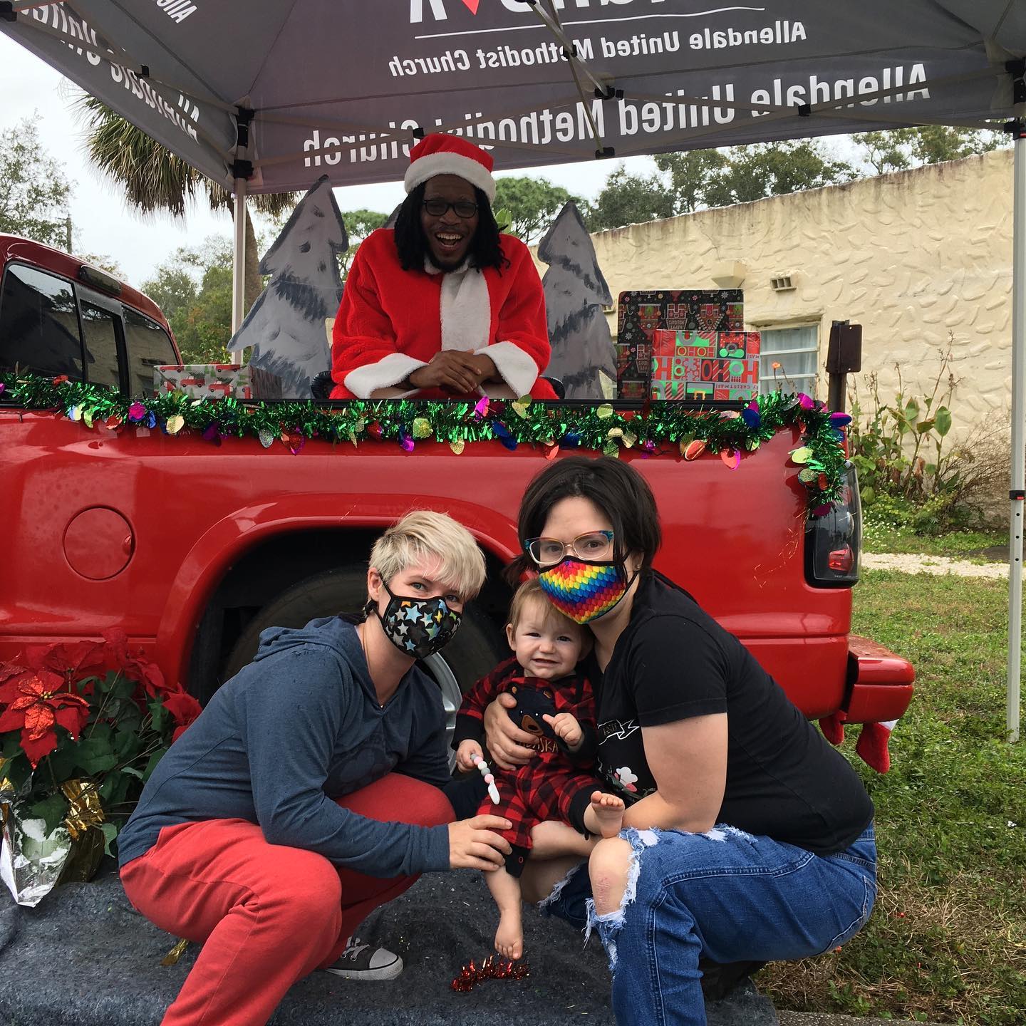 Photo of a two adults squatting and posing with their small child in front of Santa who is sitting in the back of a decorated red pickup truck with a gleeful look on his face. Decorations include presents, Christmas trees and poinsettias. They are outside with the church building in the background and a canopy tent shading the back of the truck.