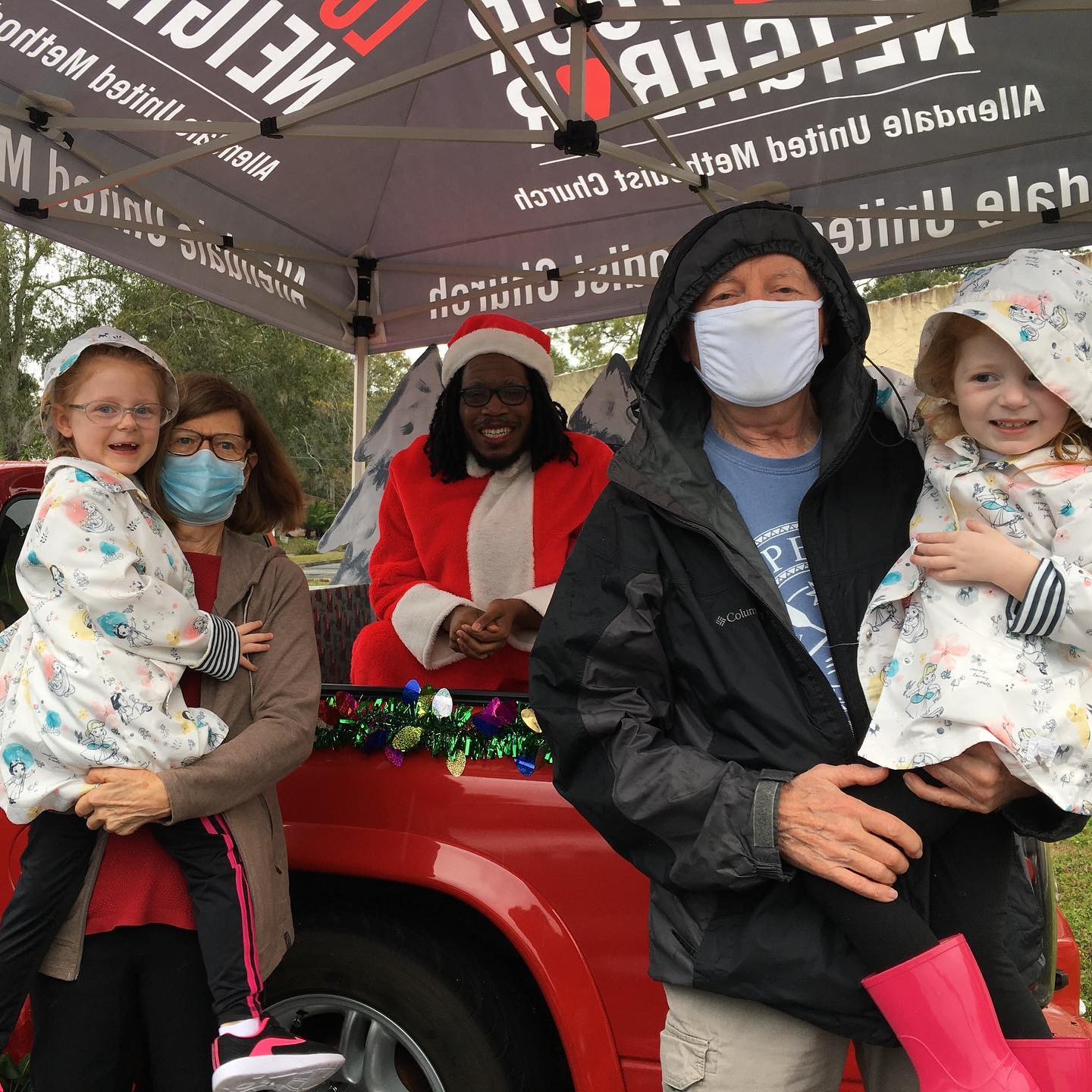 A photo of two adults, each holding a small child posing with Santa who is sitting in the back of a red pickup truck surrounded by Christmas decorations. It looks cold and rainy, but everyone is smiling gleefully. The group is sheltered from the rain by a pop up canopy tent.