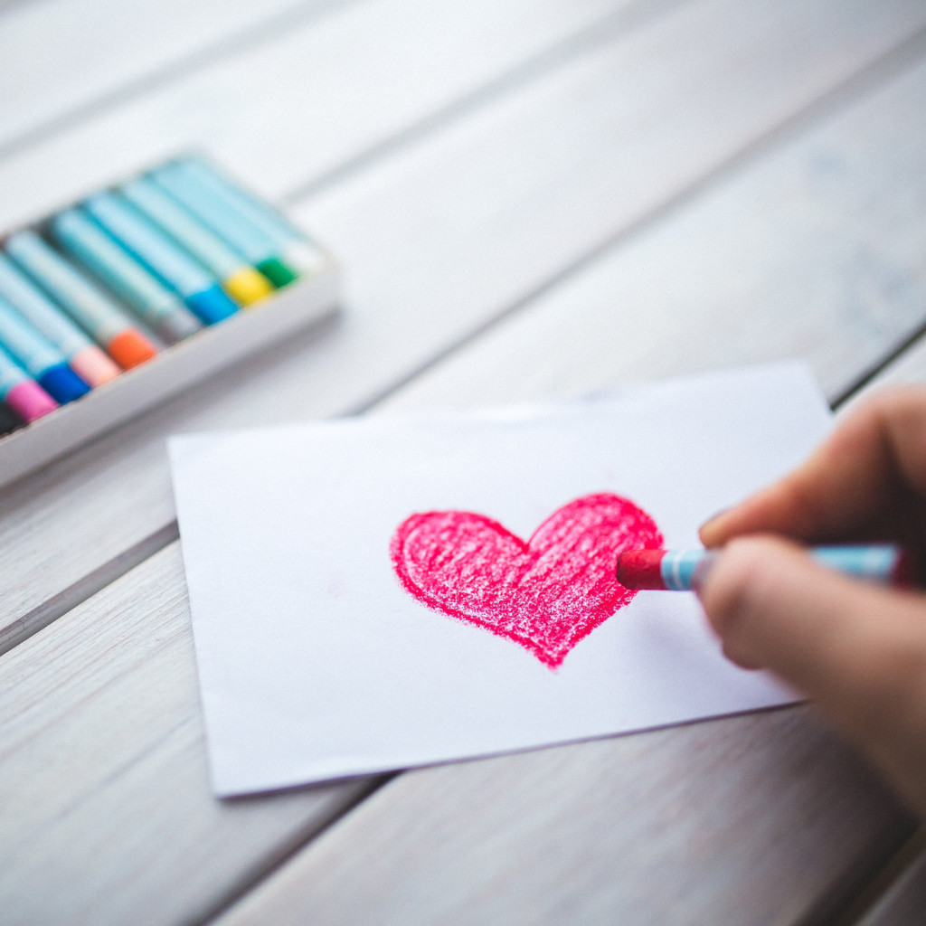 Photo of a person drawing a pink heart with a crayon on a small piece of paper.